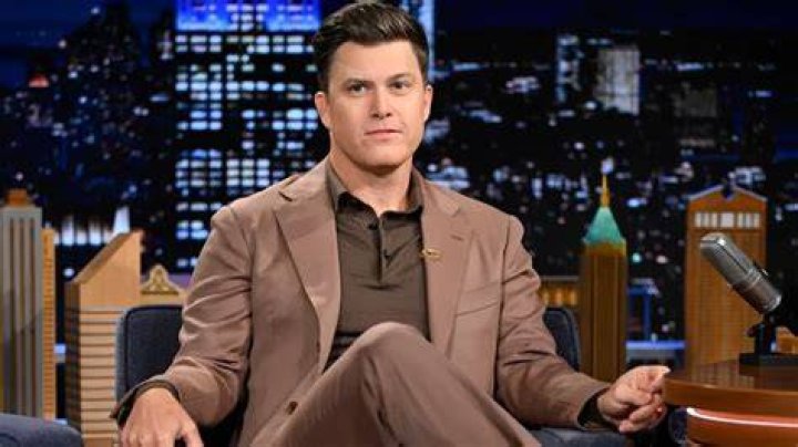 Colin Jost sitting on a chair with a mystical wand in his left hand and bookshelves behind him.